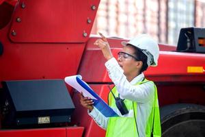 Woman Foreman in hardhat and safety vest holding holding clipboard checklist and talks on two-way radio control loading containers box from cargo photo