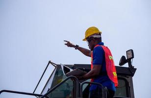 Engineer man in hard hat and safety vest holding laptop standing on container stackers control loading containers box from cargo photo