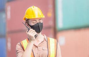 Woman Foreman in hardhat and safety vest holding holding clipboard checklist and talks on two-way radio control loading containers box from cargo photo