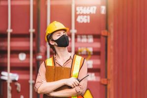 Engineer in hardhat and safety vest holding clipboard checklist and two-way radio, Woman wearing protection face mask against coronavirus at containers cargo photo