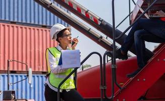 Woman Foreman in hard hat and safety vest holding clipboard checklist and talks on two-way radio, Engineer team control loading containers box from cargo photo