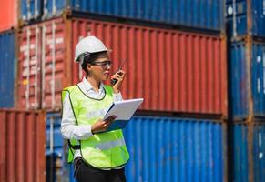 Female factory woeker in hard hat and safety vest holding clipboard checklist and talks on two-way radio control loading containers box from cargo photo