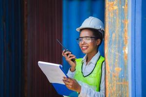 Woman Foreman in hardhat and safety vest holding clipboard checklist and talks on two-way radio control loading containers box from cargo photo