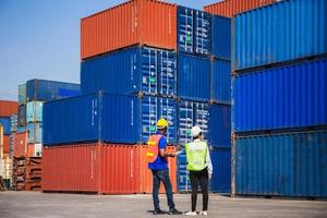 Female foreman with safety vest holding laptop and  worker man in hardhat checking and control loading containers box from cargo photo
