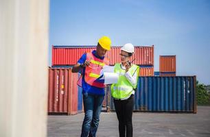 Worker man in hardhat and safety vest holding clipboard checklist and Female foreman talks on two-way radio control loading containers box from cargo photo