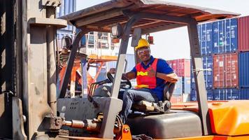 hombre trabajador con casco y chaleco de seguridad sentado en apiladores de contenedores sonriendo con los pulgares hacia arriba como señal de éxito foto