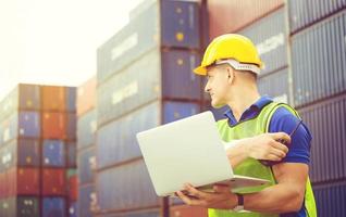 Worker man in hardhat and safety vest holding laptop and two-way radio, Foreman control loading containers box from cargo photo