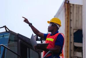 Worker man in hardhat and safety vest holding laptop standing on container stackers control loading containers box from cargo photo