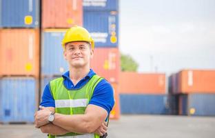 Cheerful factory worker man in hard hat smiling and arms crossed as sign of success with blurred container box background photo