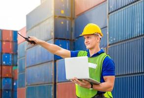 Foreman worker in hard hat and safety vest smiling and pointing control loading containers box from cargo photo