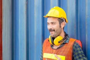 hombre trabajador de fábrica divertido en sombrero duro sonriendo a los contenedores de carga, ingeniero de felicidad en el sitio de construcción foto