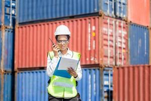 Woman Foreman in hardhat and safety vest holding holding clipboard checklist and talks on two-way radio control loading containers box from cargo photo