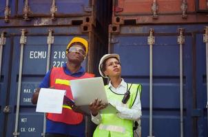 Worker man in hardhat and safety vest holding clipboard checklist and Female foreman using laptop control loading containers box from cargo photo