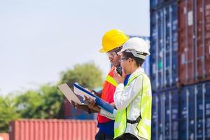 Worker man in hardhat and safety vest holding laptop and Female foreman talks on two-way radio control loading containers box from cargo photo