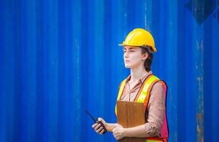 Woman Foreman in hardhat and safety vest holding holding clipboard checklist and two-way radio control loading containers box from cargo photo