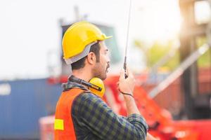 Foreman worker in hardhat and safety vest talks on two-way radio control loading containers box from cargo photo