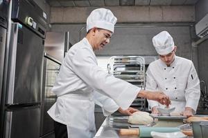 Two professional Asian male chefs in white cook uniforms and aprons are kneading pastry dough and eggs, preparing bread and fresh bakery food, baking in oven at stainless steel kitchen of restaurant. photo