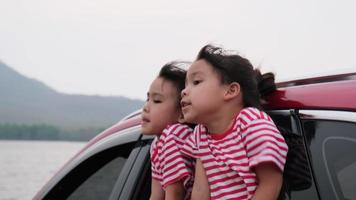 schattige aziatische broers en zussen meisjes glimlachen en hebben plezier met het reizen met de auto en kijken uit het autoraam. gelukkige familie genieten van road trip op zomervakantie. video