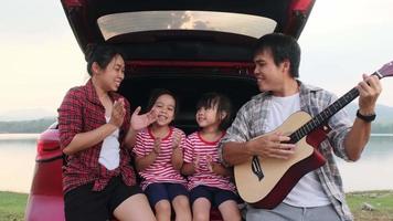 Happy family enjoying road trip on summer vacation. Mother and child sit in the trunk of the car singing along with dad playing the guitar. Holiday and travel family concept.