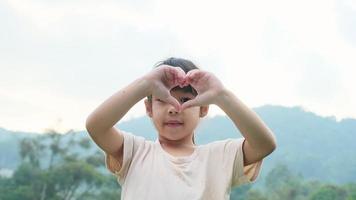 bonito pré-escolar asiático sorrindo alegremente e fazendo as mãos em forma de coração na cabeça no fundo verde da natureza. video