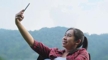 una linda mujer asiática se sienta en una silla de campamento y se toma una selfie con su teléfono inteligente durante el campamento. video
