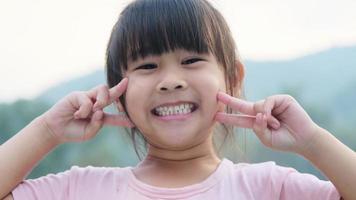 retrato de um pré-escolar asiático bonito sorrindo alegremente no jardim de verão. menina criança asiática mostrando os dentes da frente com um grande sorriso no fundo verde da natureza. video