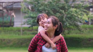 glückliche asiatische Mutter, die einem süßen kleinen Mädchen im Sommerpark eine Huckepackfahrt gibt. mutter und tochter, die im park spazieren gehen und die schöne frühlingsnatur genießen. video