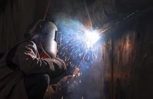 Welder in welding mask is welding metal wall of the old fishing vessel at shipyard in dark tone style photo