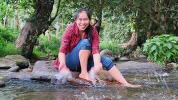 une belle femme asiatique est assise sur la pierre au milieu d'un ruisseau dans une forêt de montagne et joue avec de l'eau. concepts de vacances et d'été.