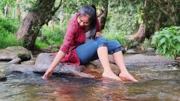 una hermosa mujer asiática se sienta en la piedra en medio de un arroyo en un bosque de montaña y juega con agua. conceptos de vacaciones y verano. video