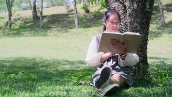 Asian woman sitting under a tree reading a book in a park.