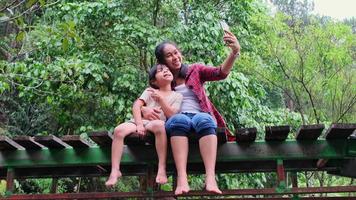 feliz madre asiática y su hija se sientan en un puente de madera a través de un arroyo en un bosque de montaña y disfrutan de la naturaleza. la familia pasa tiempo juntos en vacaciones. video
