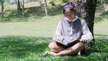 donna asiatica seduta sotto un albero che legge un libro in un parco. video