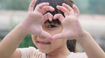 Cute Asian preschooler smiling happily making heart-shaped hands on eyes on green nature background.