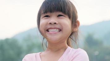 retrato de um pré-escolar asiático bonito sorrindo alegremente no jardim de verão. menina criança asiática mostrando os dentes da frente com um grande sorriso no fundo verde da natureza. video