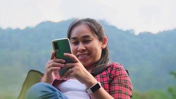mulher bonita asiática senta-se em uma cadeira de acampamento e usa um smartphone para se comunicar durante o acampamento. video