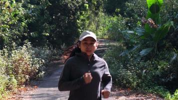 femme asiatique faisant du jogging sur un chemin de terre dans la forêt pour faire de l'exercice. femme sportive courant sur une route de campagne sous des arbres verts feuillus le matin. video