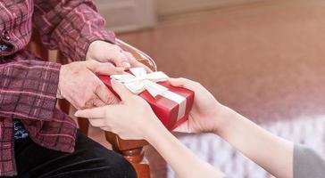 Teenage asia girl is giving grandma sitting on chair beautiful giftbox for mothers day birthday celebration idea at home with dark background, close up, copy space, cropped view photo