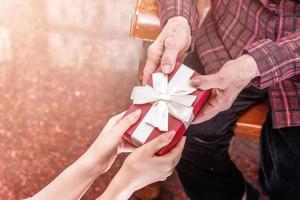 Teenage asia girl is giving grandma sitting on chair beautiful giftbox for mothers day birthday celebration idea at home with dark background, close up, copy space, cropped view photo