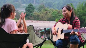 Mother and child play guitar and sing together on camping chairs near tent at camp in summer forest. The family spends time together on vacation.