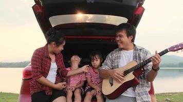 gelukkige familie genieten van road trip op zomervakantie. moeder en kind zitten in de kofferbak van de auto en zingen mee terwijl vader gitaar speelt. vakantie en reizen familie concept. video