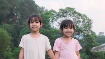 Zwei süße asiatische Schwestern, die Händchen halten, stehen im Sommergarten. Porträt von zwei süßen kleinen Mädchen, die im Park lächeln und spielen. video