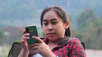 mulher bonita asiática senta-se em uma cadeira de acampamento e usa um smartphone para se comunicar durante o acampamento. video