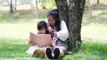 une mère et une fille asiatiques heureuses s'assoient sous un arbre sur la pelouse du jardin d'été et lisent un livre ensemble.
