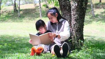 la madre e la figlia asiatiche felici si siedono sotto un albero sul prato del giardino estivo e leggono un libro insieme. video