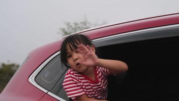 menina asiática bonitinha sorrindo e se divertindo para viajar de carro e olhando pela janela do carro. família feliz aproveitando a viagem nas férias de verão.