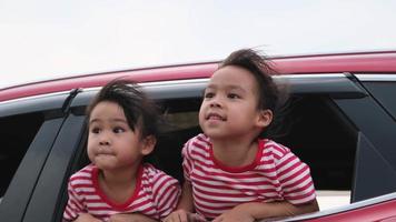 Cute Asian siblings girls smiling and having fun traveling by car and looking out of the car window. Happy family enjoying road trip on summer vacation.