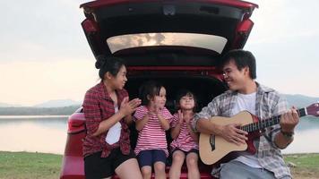 familia feliz disfrutando de un viaje por carretera en vacaciones de verano. madre e hijo se sientan en el maletero del coche cantando junto con papá tocando la guitarra. concepto familiar de vacaciones y viajes. video