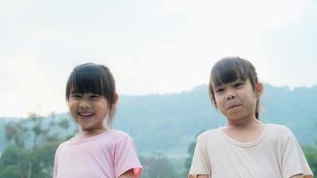retrato de dos lindas hermanas asiáticas sonriendo alegremente en el jardín de verano. niñas pequeñas asiáticas que muestran los dientes delanteros con una gran sonrisa en el fondo verde de la naturaleza. video