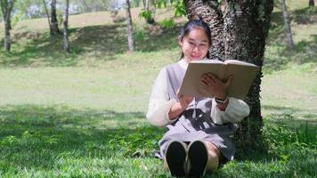 asiatisk kvinna som sitter under ett träd och tar en tupplur medan du läser en bok i en park. video